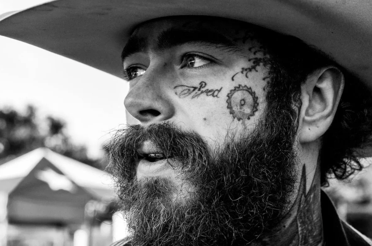 Black and white close-up portrait of musician Post Malone wearing a cowboy hat, displaying his facial tattoos and beard looking off-camera.